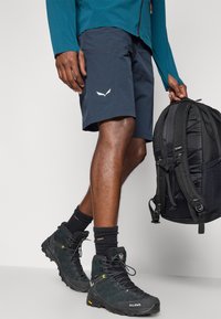 Ανδρικά ναυτικά σορτς με λευκό λογότυπο, συνδυασμένα με μαύρα ορειβατικά μποτάκια και μαύρο σακίδιο. Διαθέτουν υφασμάτινη υφή και αθλητική γραμμή.