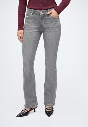 Person trägt graue Straight-Leg-Jeans, bordeauxfarbenes Langarmoberteil und schwarze spitze High Heels mit zwei Riemen. Eine Hand in der Tasche.