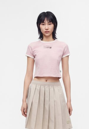 Femme aux cheveux noirs mi-longs portant un t-shirt court rose clair New Balance et une jupe plissée beige sur un fond uni.