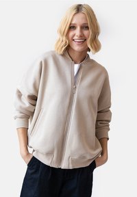 Beżowa bluza rozpinana z okrągłym dekoltem, ściągaczami na rękawach i dwiema bocznymi kieszeniami. Miękki materiał o luźnym kroju i minimalistycznym designie.