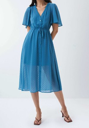 Kvinde iført en blå midikjole med flagrende ærmer, knaplukning foran, snøre i taljen og tekstureret stof, kombineret med brune sandaler med hæl.