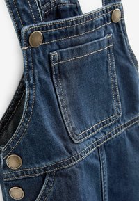 Nahaufnahme einer blauen Denim-Latzhose, die die Fronttasche, metallische Knöpfe und die detaillierte Naht auf dem Stoff zeigt.