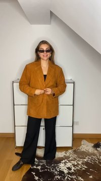 Blazer de camurça castanha com grandes lapelas, bolsos frontais e fecho de botão, combinado com calças largas pretas e sapatos escuros estampados.