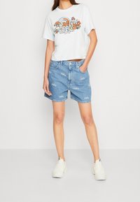 Žena v bílém grafickém crop topu s duhou a květinami, modrých džínových šortkách s bílými logy a bílých teniskách Nike.