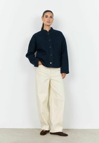 Geacă texturată bleumarin cu guler înalt, închidere cu nasturi în față și două buzunare frontale, asortată cu pantaloni largi crem și pantofi maro închis.