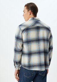 Camisa de cuadros en beige y azul marino, fabricada en un tejido suave. Presenta mangas largas, cuello abotonado y un dobladillo curvado con aberturas laterales.