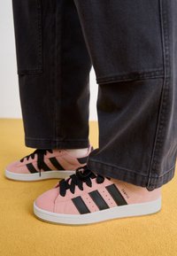 Růžové semišové tenisky s černými pruhy a tkaničkami, s bílou podešví. Jemná textura a moderní design. Nosené s tmavými kalhotami na žluté podlaze.