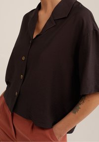 Camisa negra de manga corta, con collar y corte relajado, que presenta una tela texturada y tres grandes botones redondos en la parte frontal.
