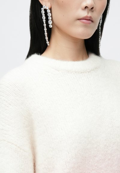Lange krystal øreringe med et lineært design, der har klare sten indsat i et sølvfarvet metal, parret med en blød, hvid strikket sweater.