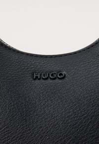 Materiale in pelle nera con superficie testurizzata. Presenta un'apertura arrotondata e il logo "HUGO" in rilievo con finitura lucida.