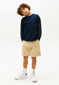 Maglietta a maniche lunghe blu navy con striscia orizzontale, pantaloni corti beige chiaro e sneakers bianche. Presenta un logo sulla manica della maglietta e dettagli in tessuto.
