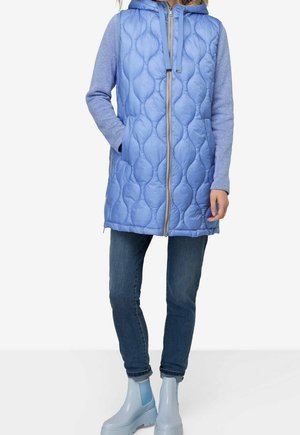 Kvinde iført en lyseblå quiltet ærmeløs vest over en blå hættetrøje, mørke jeans og lyseblå ankelstøvler, står med den ene hånd i lommen.