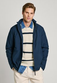 Marinblå pufferjacka med dragkedja och huva, bärs över en krämfärgad stickad tröja med marinblå ränder och en denimskjorta med krage under.