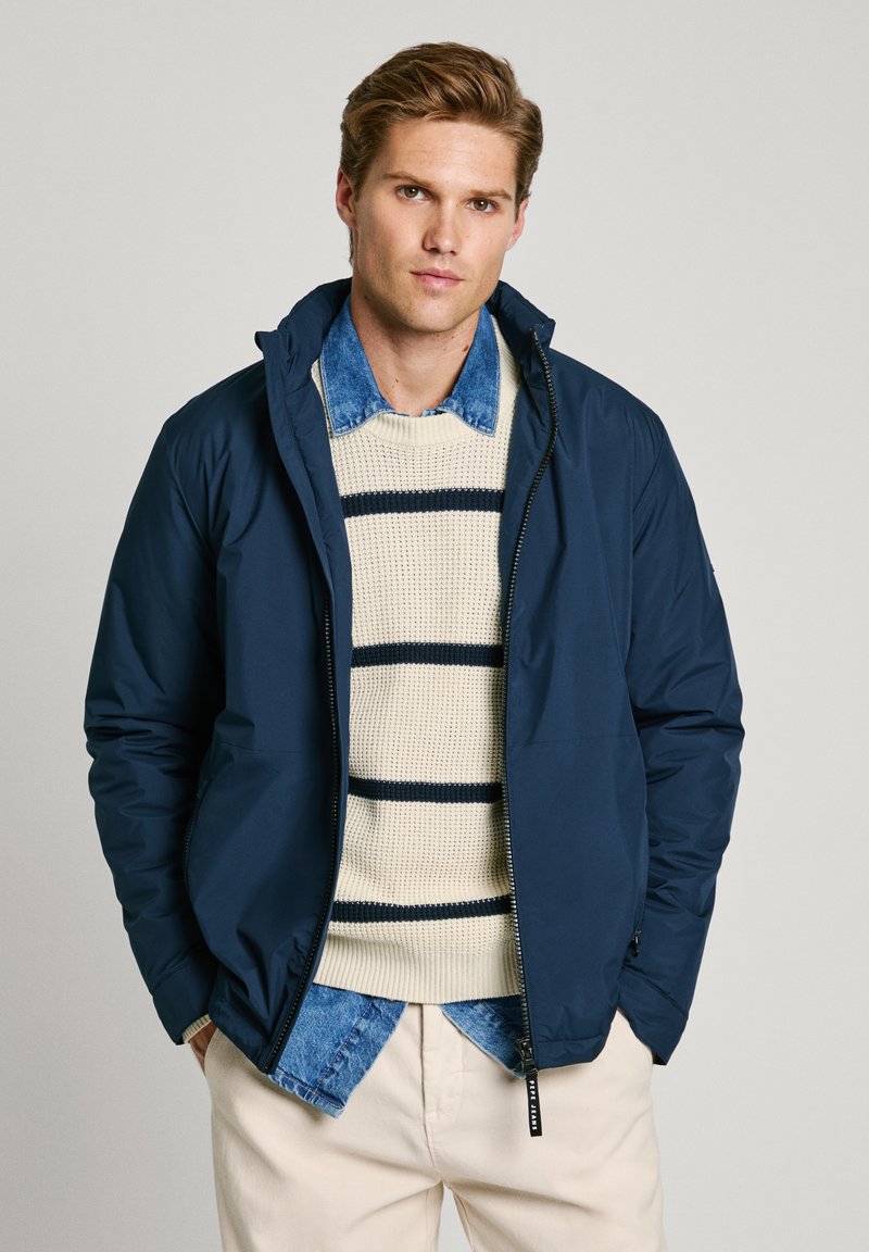 Marinblå pufferjacka med dragkedja och huva, bärs över en krämfärgad stickad tröja med marinblå ränder och en denimskjorta med krage under.