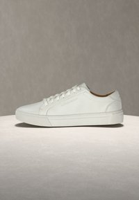 Λευκό δερμάτινο χαμηλό sneaker με κορδόνια σε ουδέτερη επιφάνεια και μονόχρωμο γκρι φόντο, εμφανιζόμενο από την εξωτερική πλευρά.