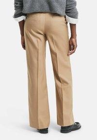 Persona che indossa pantaloni beige a gamba larga, maglione grigio con maniche arrotolate e mocassini neri, in piedi contro uno sfondo bianco.