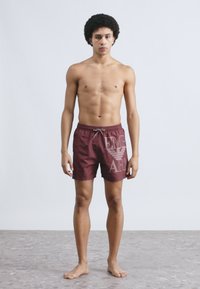 Bordó úszónadrág logónyomattal, elasztikus derékrésszel és húzózsinórral. Könnyű anyag, sima tapintású és térd fölé érő szabású.