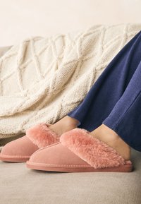 Chaussons en daim rose sans lacets avec une tige en peluche rose, présentant des coutures décoratives et une semelle durable, posés sur une surface en tissu clair.