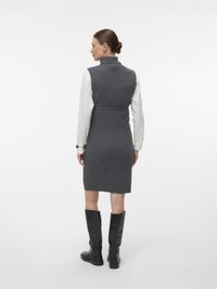 Szürke bordázott kötött ruha magas gallérral, testhez simuló derékkal és térdig érő hosszúsággal. Fekete magas csizmával párosítva, egyszerű háttér előtt.