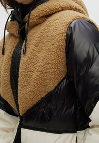Giacca a vento con cappuccio in finta shearling color sabbia e corpo in sintetico nero; presenta cordoncini regolabili e pannelli a contrasto.