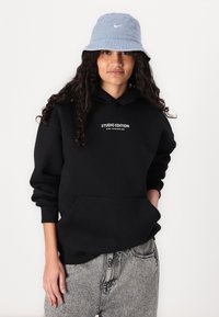Čierna mikina s kapucňou so žltým nápisom "STUDIO EDITION", v kombinácii s modrým vedrom s bielym logom Nike. Neformálne šedé džínsy s poškodením.