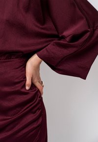 Bordeaux jurk met textuurstof, gedrapeerd ontwerp, lange mouwen en geplooide details bij de taille. Hand op de heup geplaatst.