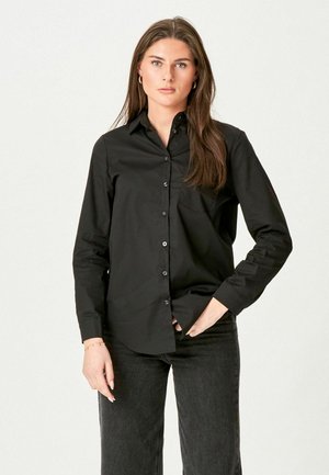 Frau mit langen braunen Haaren, die eine schwarze Hemdbluse und dunkle Jeans trägt, steht mit einer Hand in der Tasche vor einem schlichten Hintergrund.