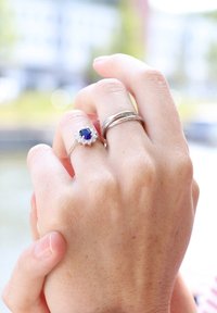 Zilveren ringen op een hand, waarvan één met een centrale blauwe steen omringd door kleinere stenen, en de ander met een gedraaide bandontwerp. Rechte huidtextuur.