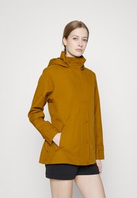 Casaco de chuva amarelo mostarda com capuz, fechos de pressão na frente, bolsos laterais e punhos ajustáveis, feito de um material liso e resistente à água.