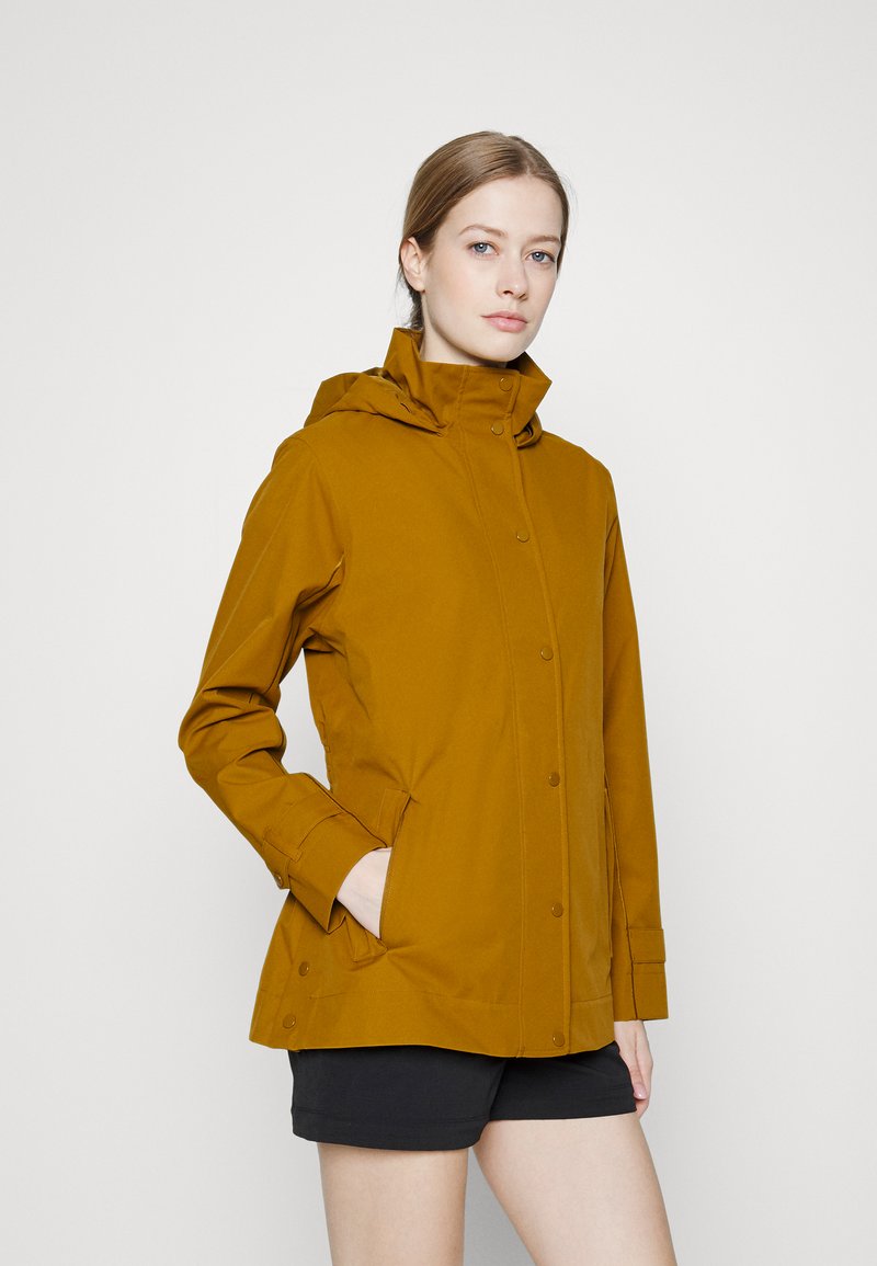 Casaco de chuva amarelo mostarda com capuz, fechos de pressão na frente, bolsos laterais e punhos ajustáveis, feito de um material liso e resistente à água.