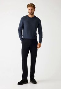 Marineblauer Pullover mit Rautenmuster, kombiniert mit schwarzer Hose und schwarzem Schuhwerk; lässige Passform, der Stoff wirkt weich und strukturiert.