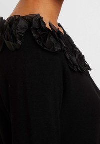 Svart tröja med en åtsittande silhuett. Texturerad, volangkantad svart tyg framhäver halsringningen och ger dimension. Mjuk stickad material.