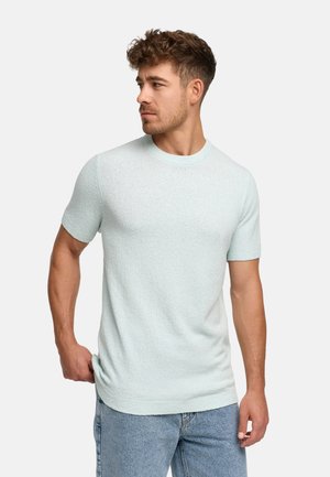 Mand med kort krøllet hår iført lyseblå kortærmet striktrøje og lyseblå jeans, kigger mod venstre med hånden i lommen.