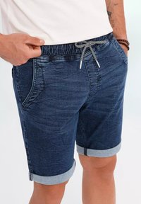 Pantalones cortos de mezclilla azul con cinturilla elástica, cordón ajustable y puños enrollados. Presenta bolsillos delanteros y una textura suave.