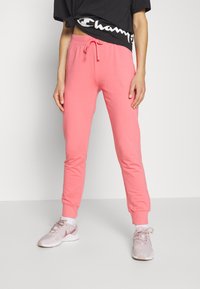 Rosa Jogginghosen mit einem Kordelzugbund, Bündchen an den Knöcheln und einer weichen Textur, kombiniert mit neutral gefärbten Sneakern und einem schwarzen Cropped-Shirt.