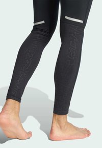 Čierne športové legíny s textúrovaným vzorom, s reflexnými akcentami na stehnách a tesným strihom, ukazujúce nahú nohu.