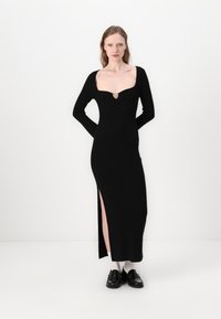 Abito lungo a maniche lunghe nero a coste con scollatura quadrata, caratterizzato da un'accenno a forma di cuore sul petto e uno spacco laterale all'orlo.