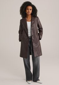 Trench de piel marrón con una silueta larga, solapas con muesca, botones frontales y bolsillos laterales. Combinado con unos pantalones vaqueros anchos y zapatos de puntera.