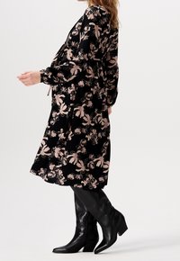 Musta kukkamekko, jossa on vaaleanpunaisia kukkakuvioita, pitkät hihat ja rento leikkaus. Yhdistetty polven ylle ulottuviin mustiin saappaisiin, joissa on terävä nenä.