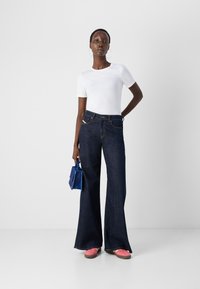 Femme portant une chemise blanche à manches courtes, un jean large bleu foncé, des chaussures roses, tenant un petit sac à main bleu, debout devant un fond blanc uni.