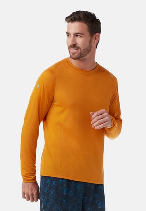 Lange mouw shirt gemaakt van lichtgewicht oranje stof met een ronde halslijn en raglan mouwen, gedragen met gebloemde blauwe shorts.