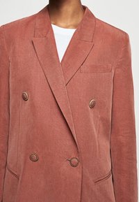 Blazer doppiamente abbottonato in un colore rosa polveroso, realizzato in tessuto liscio. Presenta rever a punta, una tasca sul petto e bottoni rotondi in tono oro.