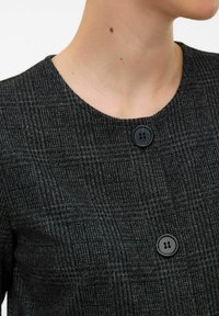 Närbild av en person klädd i en mörkgrå rutig jacka med stora svarta knappar och rund halsringning, som visar en del av nacken och hakan.