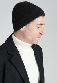 Schwarze gerippte Strickmütze, getragen mit einem weißen Rollkragenpullover und einem schwarzen Mantel. Verfügt über einen engen Sitz und ein strukturiertes, vertikales Muster.