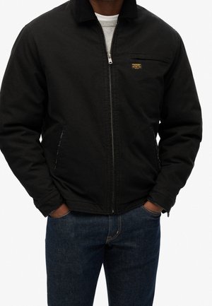 Man draagt een zwart jack met rits, kraag, voorzakken en een klein geel logo op de borst, gecombineerd met donkerblauwe spijkerbroek.