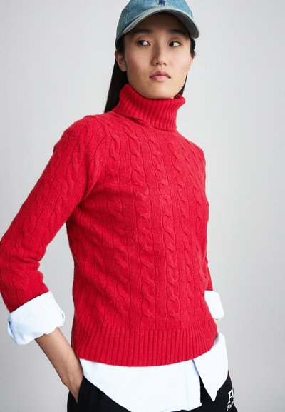 Roter, grob gestrickter Pullover mit Stehkragen und geripptem Saum. Getragen über einem weißen Hemd mit Kragen und kombiniert mit einer Jeansmütze.