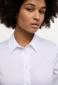 Camicia bianca con colletto classico, texture liscia e bottoni trasparenti. Primo piano sul collo e sulla parte superiore del torace.
