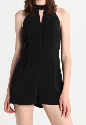 Kobieta w czarnym kombinezonie bez rękawów z wysokim dekoltem i rozcięciem z przodu, stojąca na tle jednolitego, jasnego tła.