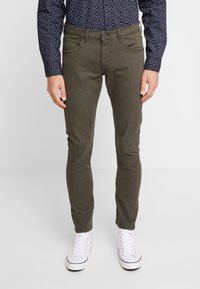 Hombre con pantalones ajustados de color verde oliva, camisa azul marino con estampado y zapatillas blancas altas, posando sobre un fondo blanco.