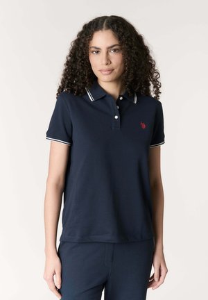 Donna con capelli ricci che indossa una polo blu navy con bordi bianchi e un piccolo logo rosso, in piedi davanti a uno sfondo semplice.